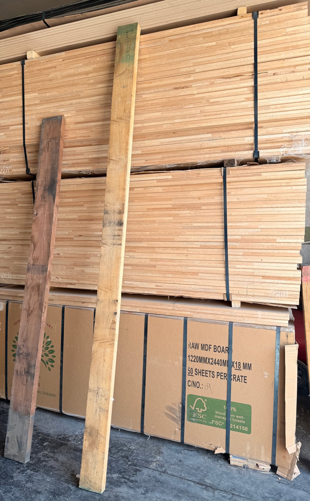A rough dark board and a rough light board leaning against stacked sheets of MDF in a warehouse