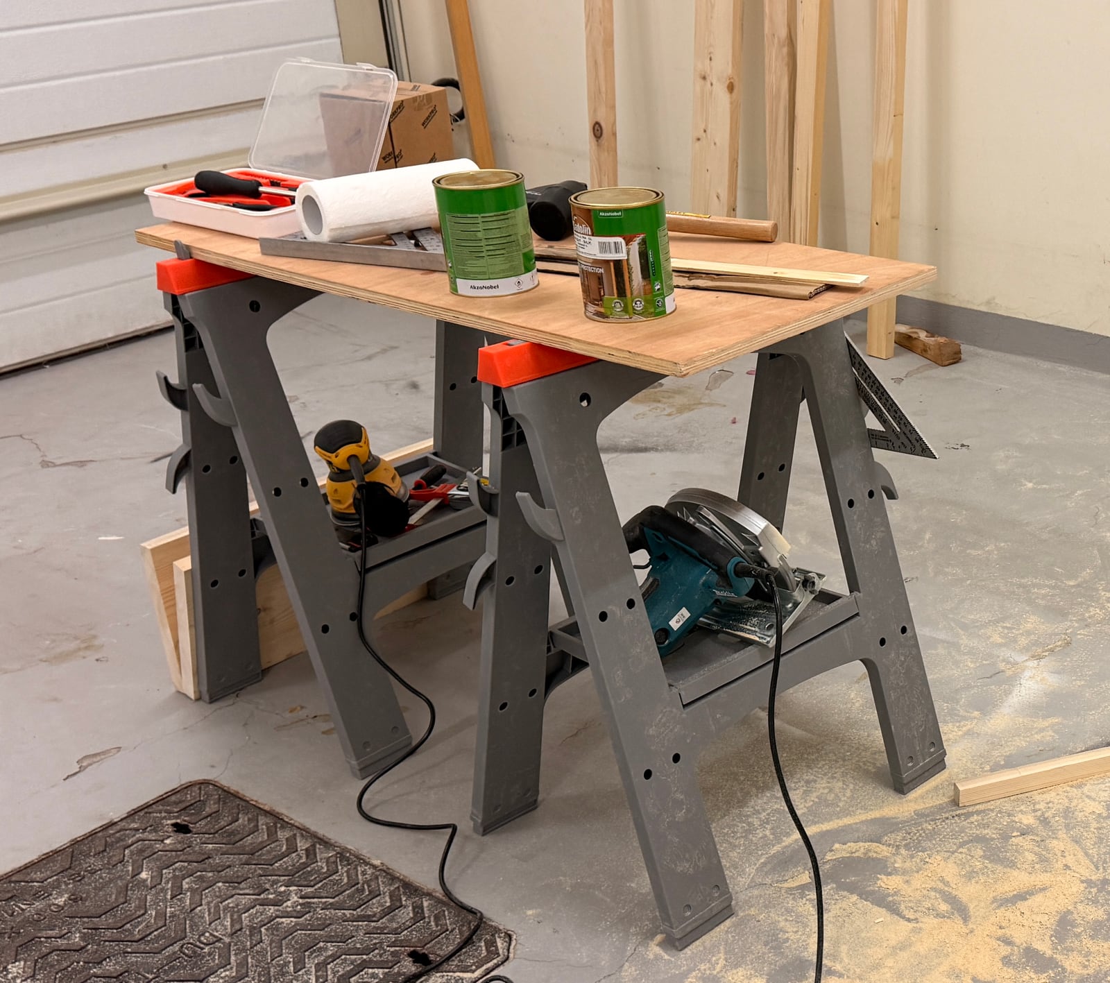 A sheet of plywood on two grey plastic sawhorses in a garage, with a circular saw and an orbital sander on the sawhorse shelves and two tins of finish on top
