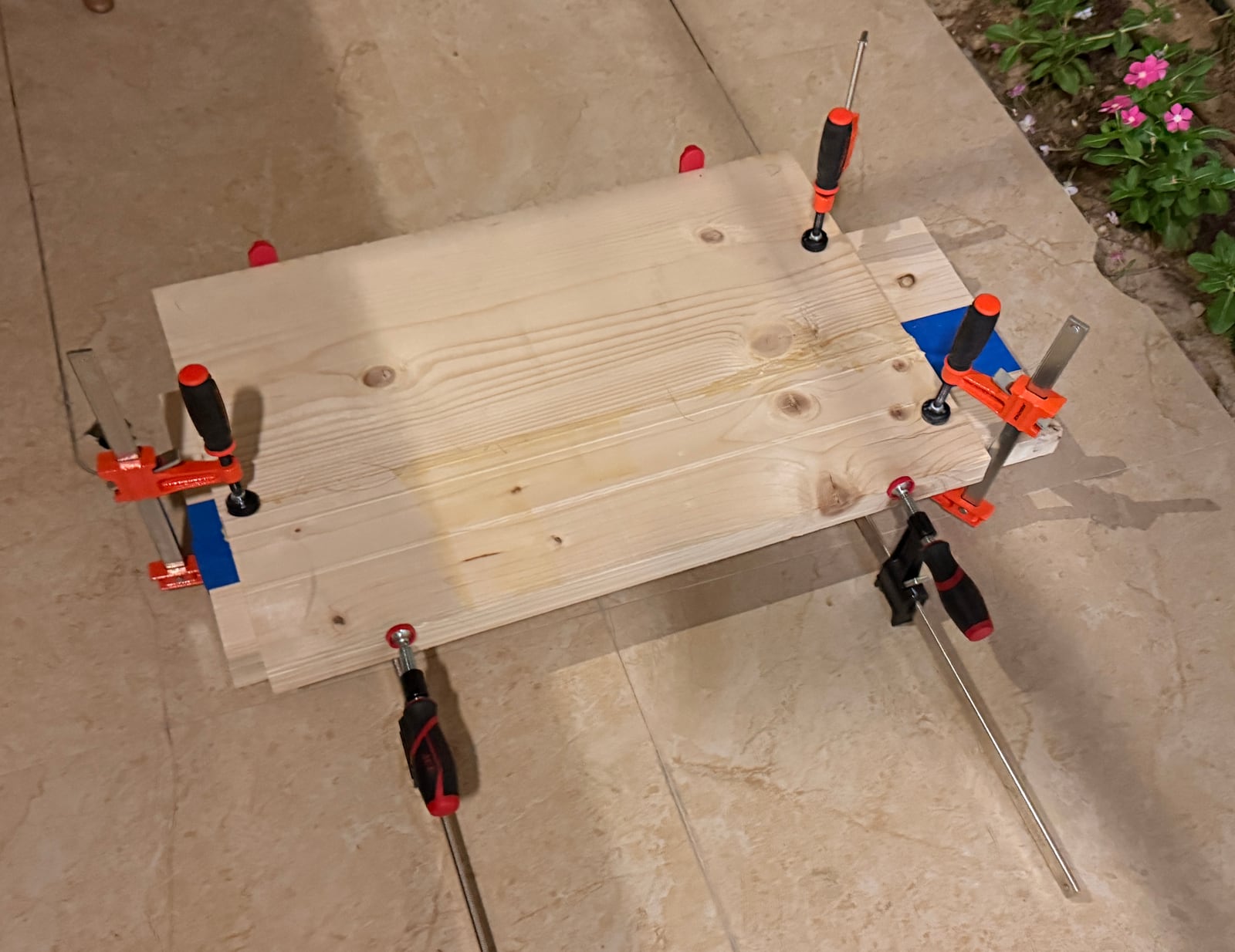 Two softwood boards glued edge to edge and held with orange F-clamps on a tiled floor