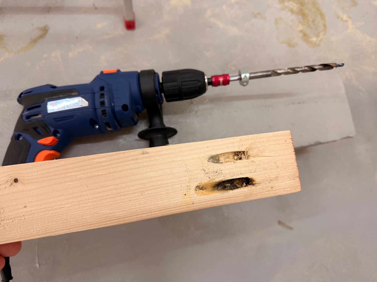 Two scorched, shallow pocket holes in a softwood board, next to a blue corded drill fitted with a pocket hole bit