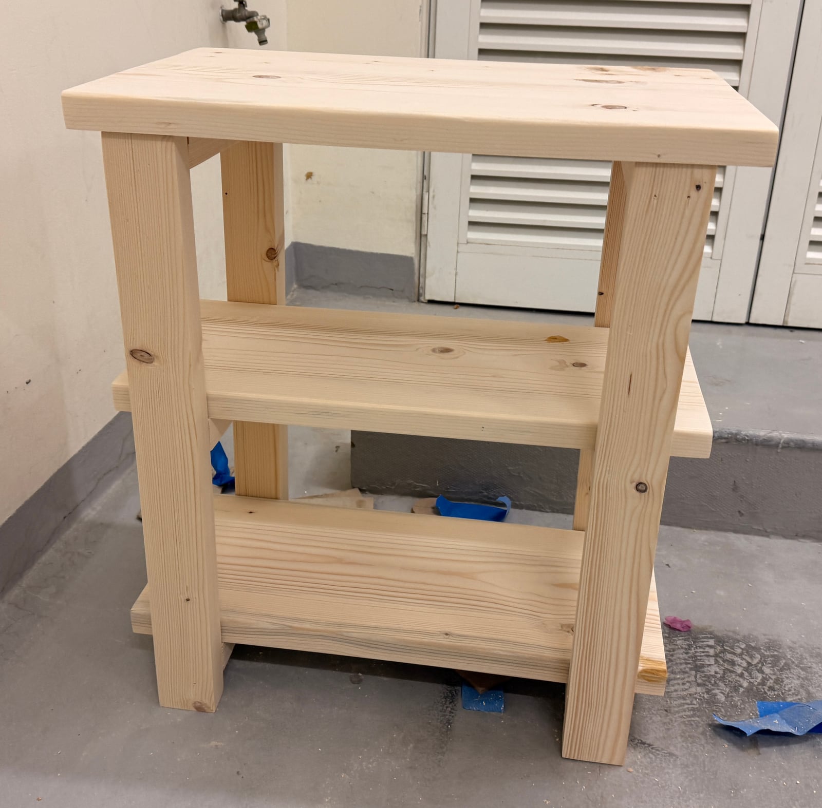 The assembled side table in bare wood: four square legs, a glued-up top and two lower shelves, standing in the garage