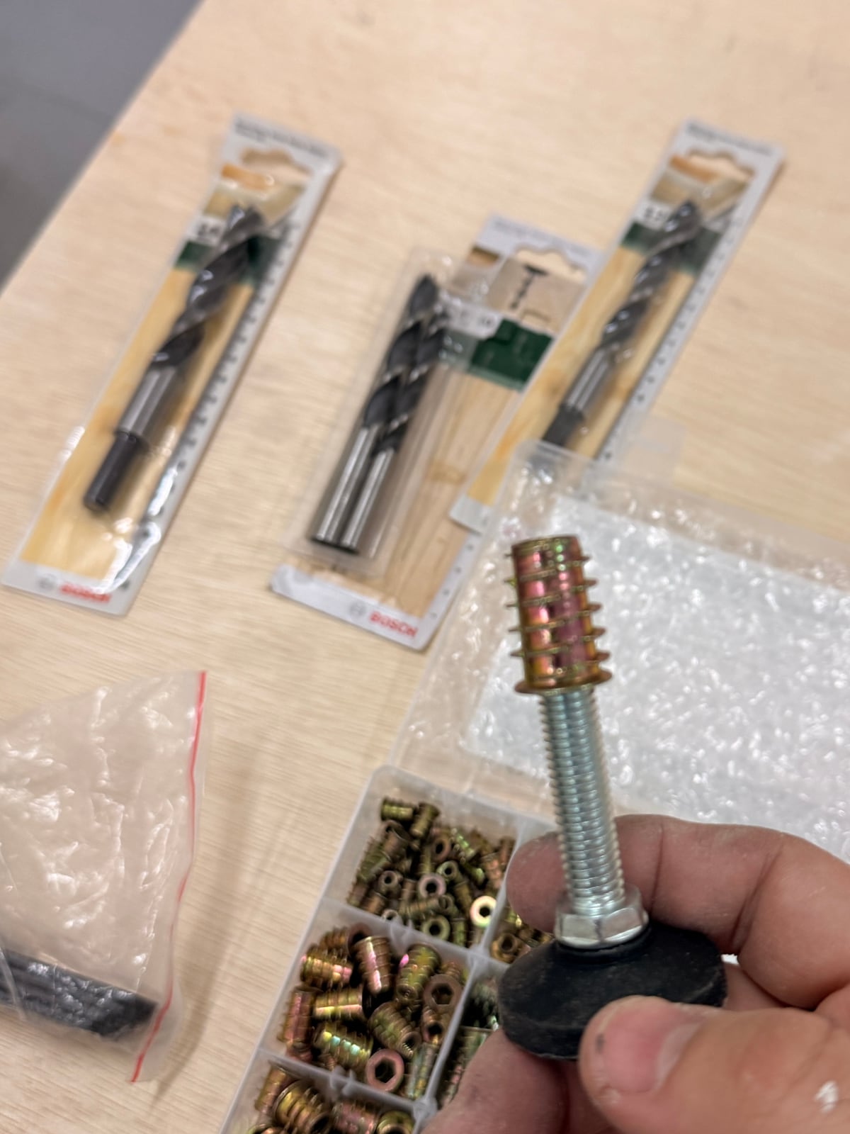 A hand holding an adjustable foot above a box of threaded inserts and a set of drill bits